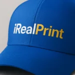 Șapcă albastră personalizată cu nume și text iRealPrint, imprimare premium alb și auriu pe fundal intens