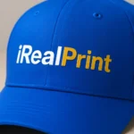Șapcă albastră personalizată cu nume iRealPrint, text alb și auriu aplicat prin transfer termic pe fundal intens