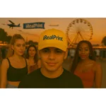 Șapcă galbenă personalizată iRealPrint – model lifestyle la festival, fundal cu roată panoramică și atmosferă vibrantă
