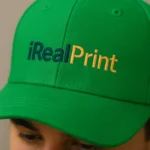 Șapcă verde personalizată cu nume iRealPrint – logo imprimat alb și galben pe material verde, detaliu apropiat al personalizării