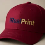 Șapcă vișinie personalizată cu text – detaliu logo iRealPrint, imprimeu auriu și albastru pe fundal intens