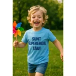 Copil purtând tricou personalizat albastru azur cu mesajul „Sunt supereroul tău!”, zâmbind și alergând prin iarbă – imagine veselă, energie pozitivă și libertate copilărească