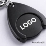 Detaliu macro breloc promoțional negru cu fisă pentru cărucior, personalizare clară tip logo, accent pe textură mată și imagine profesională de brand