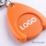 Detaliu macro breloc promoțional portocaliu cu fisă pentru cărucior, personalizare clară tip logo, accent pe textură, contrast și vizibilitate în branding promoțional
