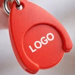 Detaliu macro breloc promoțional roșu cu fisă pentru cărucior, personalizare clară tip logo, accent pe textură și vizibilitate în branding promoțional