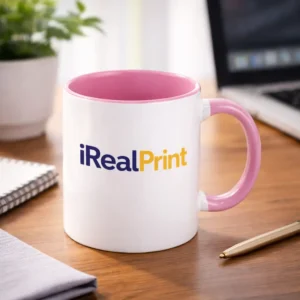 Cană personalizată cu interior roz pastel și logo iRealPrint plasată pe birou într-un cadru de lucru modern, sugerând utilizarea zilnică în spații office
