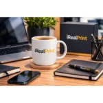 Cană albă personalizată 180ml cu logo iRealPrint într-un setup premium de birou cu agendă și pix personalizat, sugerând imagine de brand profesională
