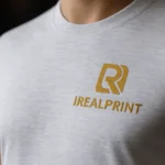 Detaliu tricou unisex personalizat gri deschis cu logo iRealPrint aplicat prin transfer termic, evidențiind textura materialului și claritatea imprimării