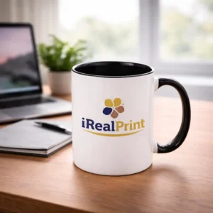 Cană personalizată cu interior negru onyx și logo iRealPrint plasată pe birou lângă laptop și agendă, sugerând utilizarea zilnică în mediul office