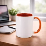 Cană personalizată cu interior portocaliu plasată pe birou lângă laptop și agendă, sugerând utilizarea zilnică într-un mediu office modern