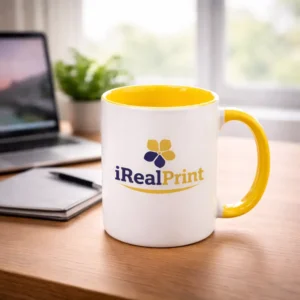 Cană personalizată cu interior galben gold și logo iRealPrint plasată pe birou lângă laptop și agendă, sugerând utilizarea zilnică într-un mediu office modern
