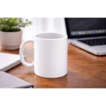 Cană albă personalizată 350ml plasată pe birou lângă laptop și agendă, sugerând utilizarea zilnică într-un mediu office modern