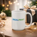 Cană albă personalizată 450ml cu logo iRealPrint prezentată lângă o cutie cadou elegantă pe fundal festiv, sugerând un cadou premium pentru ocazii speciale