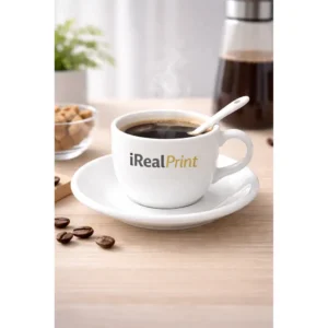 Set ceașcă cafea personalizată cu linguriță și farfurie și logo iRealPrint servit cu cafea fierbinte pe masă, alături de boabe de cafea și accesorii pentru preparare