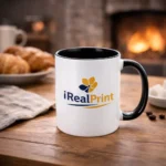 Cană personalizată cu interior negru onyx și logo iRealPrint într-un decor cozy lângă șemineu și croissant, sugerând un moment relaxant de cafea
