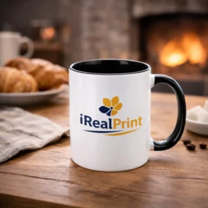 Cană personalizată cu interior negru onyx și logo iRealPrint într-un decor cozy lângă șemineu și croissant, sugerând un moment relaxant de cafea