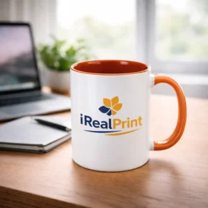 Cană personalizată cu interior portocaliu și logo iRealPrint pe birou într-un cadru luminos, evidențiind contrastul vizual și impactul personalizării