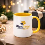 Cană personalizată cu interior galben gold și logo iRealPrint într-un decor festiv cu cutie cadou și lumini calde, sugerând ideea de cadou memorabil
