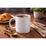 Cană albă personalizată 250ml cu cafea fierbinte într-un decor cozy cu croissant, linguriță aurie și boabe de cafea, sugerând un moment relaxant de dimineață