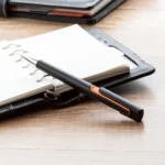 Pix plastic personalizat Nuba Black portocaliu așezat lângă agendă în context business, produs promoțional utilizat în birouri și întâlniri profesionale