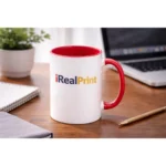 Cană personalizată cu interior roșu cherry și logo iRealPrint plasată pe birou lângă laptop și agendă, sugerând utilizarea zilnică în mediul office