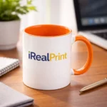 Cană personalizată cu interior portocaliu și logo iRealPrint plasată pe birou lângă laptop și agendă, sugerând utilizarea zilnică în mediul office
