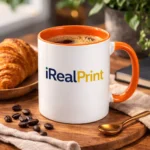 Cană personalizată cu interior portocaliu iRealPrint umplută cu cafea lângă croissant și boabe de cafea, sugerând un moment relaxant de mic dejun