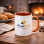 Cană personalizată cu interior portocaliu și logo iRealPrint într-un decor cozy lângă șemineu și produse de patiserie, sugerând un moment relaxant de cafea