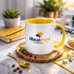 Cană personalizată cu interior galben gold și logo iRealPrint prezentată într-un decor premium cu linguriță aurie și boabe de cafea, evidențiind eleganța produsului