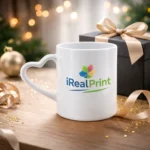 Cană personalizată 350ml cu mâner inimă și logo iRealPrint prezentată lângă o cutie cadou elegantă și fundal luminos, sugerând un cadou romantic special