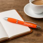 Pix plastic personalizat Elipse portocaliu așezat pe notebook pe birou lângă o ceașcă de cafea, pix promoțional modern pentru branding profesional și utilizare zilnică