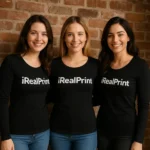 Grup de femei purtând bluze negre personalizate cu logo iRealPrint în fața unui perete din cărămidă, imagine de echipă și branding vizibil