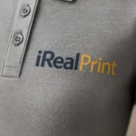 Detaliu logo iRealPrint aplicat prin transfer termic pe tricou polo damă gri închis, evidențiind textura materialului și contrastul personalizării
