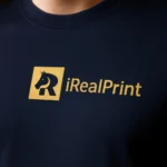 Detaliu tricou unisex personalizat negru cu logo iRealPrint aplicat prin transfer termic auriu, evidențiind textura materialului și claritatea imprimării