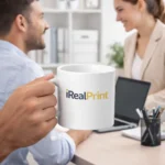 Cană albă personalizată 180ml cu logo iRealPrint într-un cadru office cu două persoane în discuție, sugerând utilizarea în mediul profesional