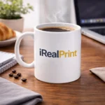 Cană albă personalizată 350ml cu logo iRealPrint pe birou lângă laptop și cafea, sugerând utilizarea zilnică într-un workspace modern