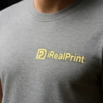 Detaliu tricou unisex personalizat gri închis cu logo iRealPrint aplicat prin transfer termic, evidențiind claritatea imprimării și textura materialului
