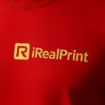 Detaliu close-up cu logo iRealPrint imprimat prin transfer termic pe tricou unisex roșu, evidențiind claritatea personalizării