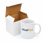 Cană albă personalizată 350ml cu logo iRealPrint prezentată lângă cutia de ambalaj pe fundal alb, evidențiind produsul pregătit pentru livrare sau cadou