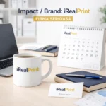 Cană albă personalizată 180ml cu logo iRealPrint pe birou lângă laptop, calendar și cărți, sugerând utilizarea ca produs promoțional de branding